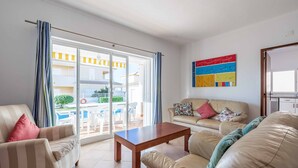 A living room with a sofa, armchair, and coffee table, a sliding glass door leading to an outdoor area, and a colorful abstract painting on the wall.