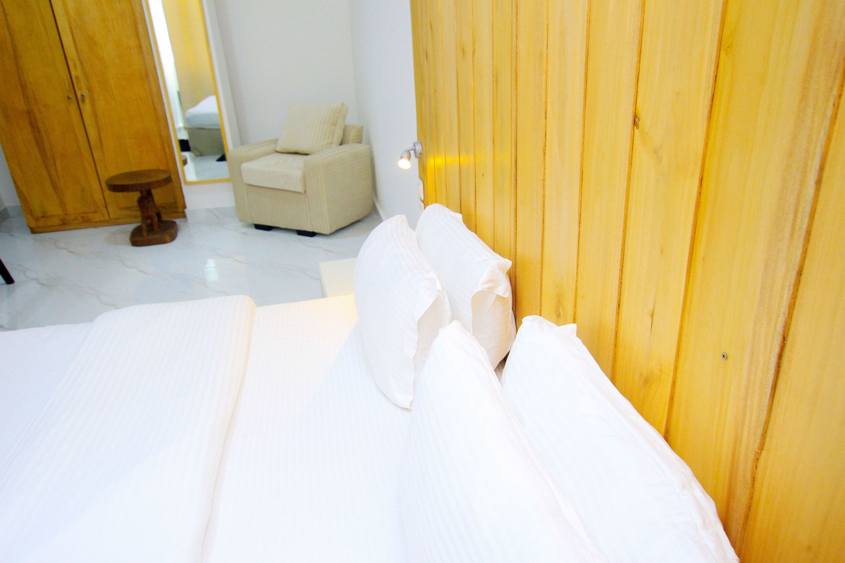 A neatly made bed with white linens, a wooden wardrobe, and a small wooden stool in a bright room with white walls and a tiled floor.