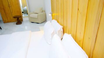 A neatly made bed with white linens, a wooden wardrobe, and a small wooden stool in a bright room with white walls and a tiled floor.