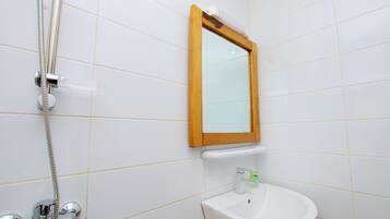 Un baño con paredes de azulejos blancos, un espejo con marco de madera, un lavabo blanco y una ducha con manguera de cromo.