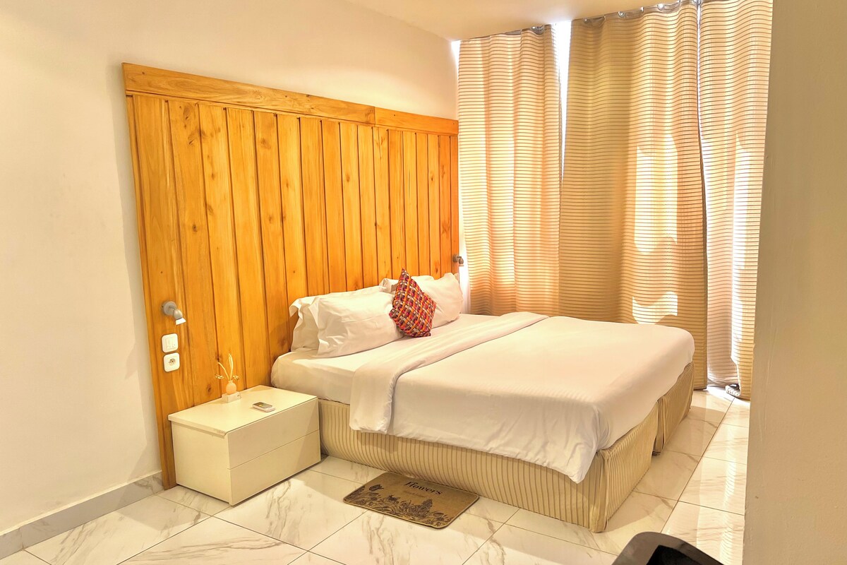 Una habitación de hotel con cabecera de madera, una cama con ropa blanca, una mesita de noche con lámpara y una ventana con cortinas a rayas.