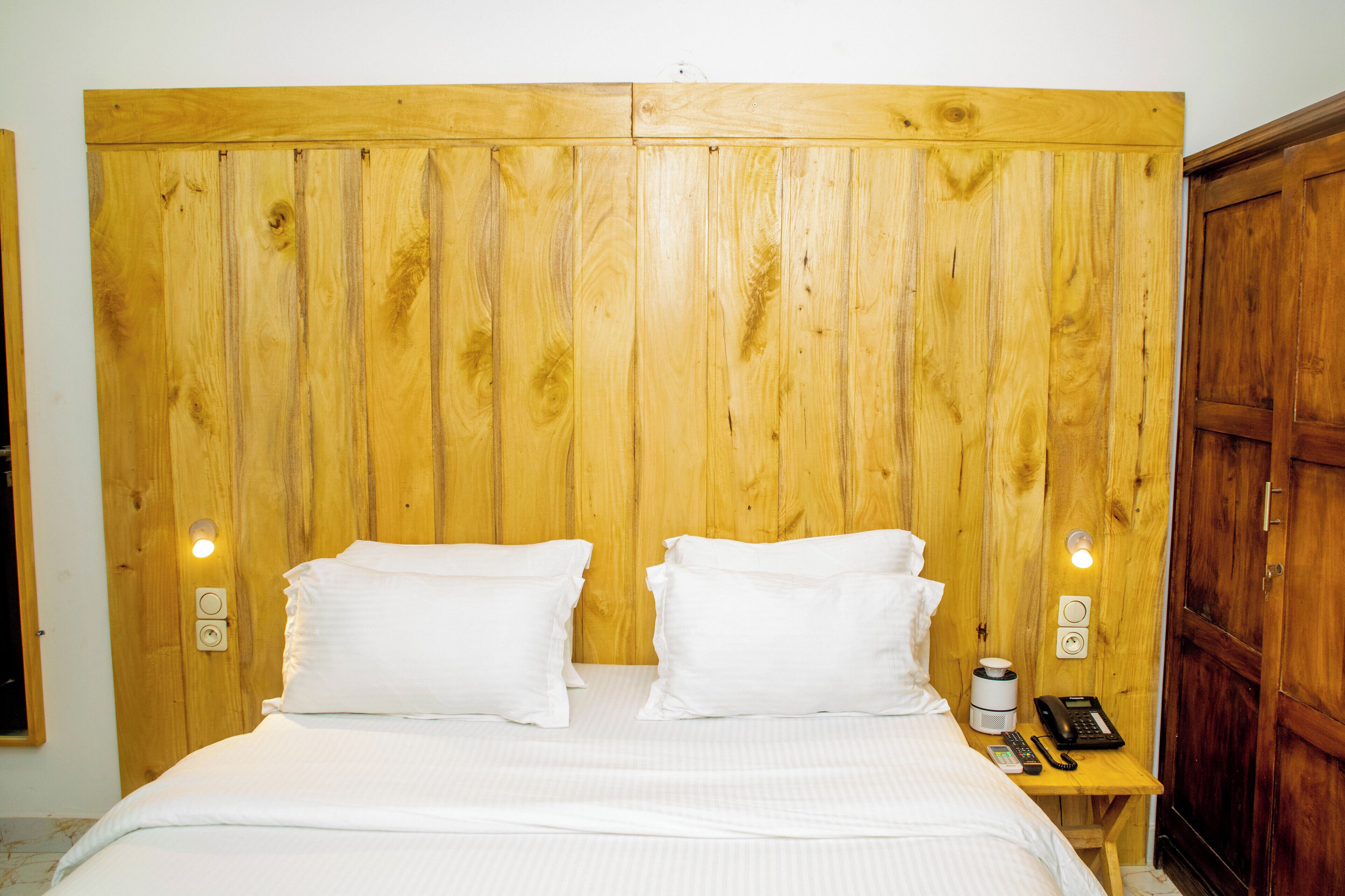 Cama con cabecera de madera, dos almohadas, una mesita de noche con teléfono y cafetera, y un armario de madera.