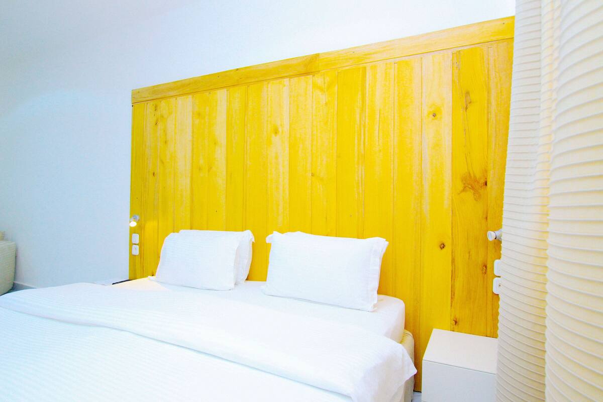 A bed with white linens, a yellow headboard, and a white nightstand.