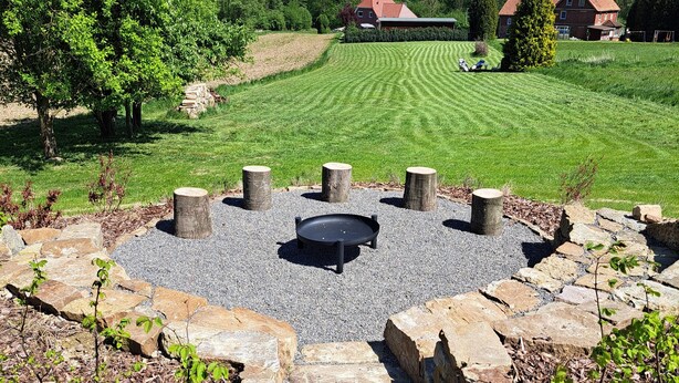 A stone fire pit surrounded by a gravel area, with a well-manicured lawn and a distant view of a residential area.