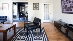 Een ruime woonkamer met een zwarte lederen fauteuil, een houten salontafel en een zwart-wit vloerkleed. Op de achtergrond is een zithoek met een tafel en stoelen te zien.