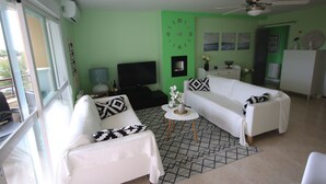 Un salon avec deux canapés blancs, un téléviseur, une horloge et un ventilateur de plafond.
