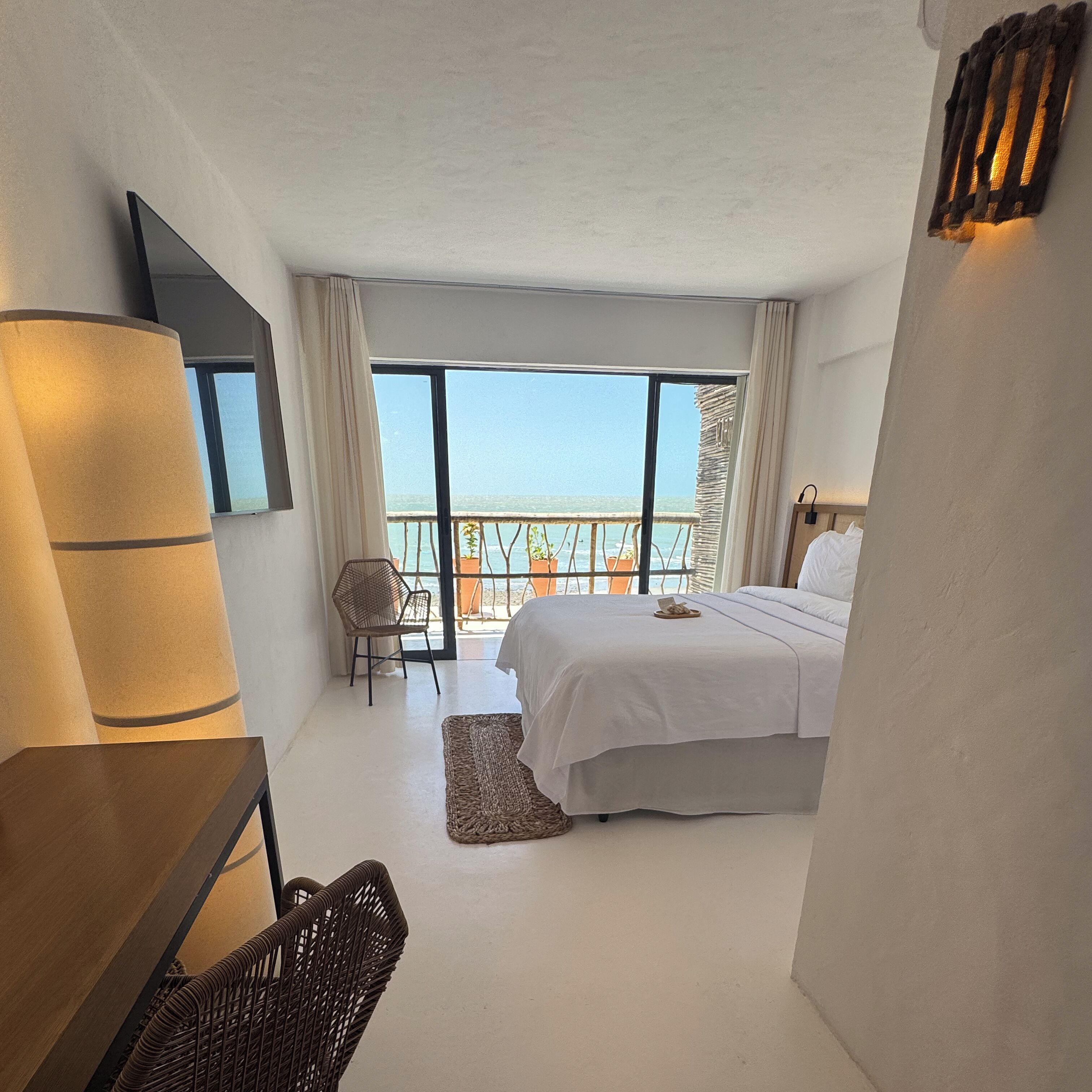Habitación de hotel con cama, silla, escritorio y un amplio ventanal con vista al mar.