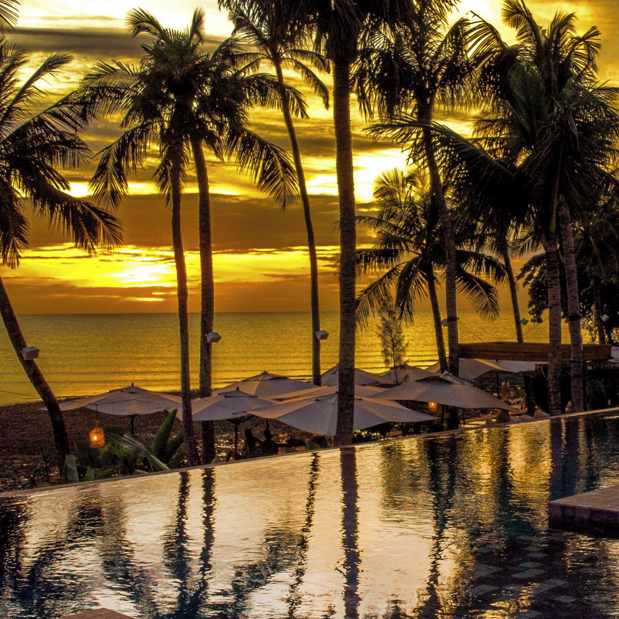 Ein Pool ohne Ende mit Palmen und Sonnenschirmen, der auf den Ozean bei Sonnenuntergang blickt.