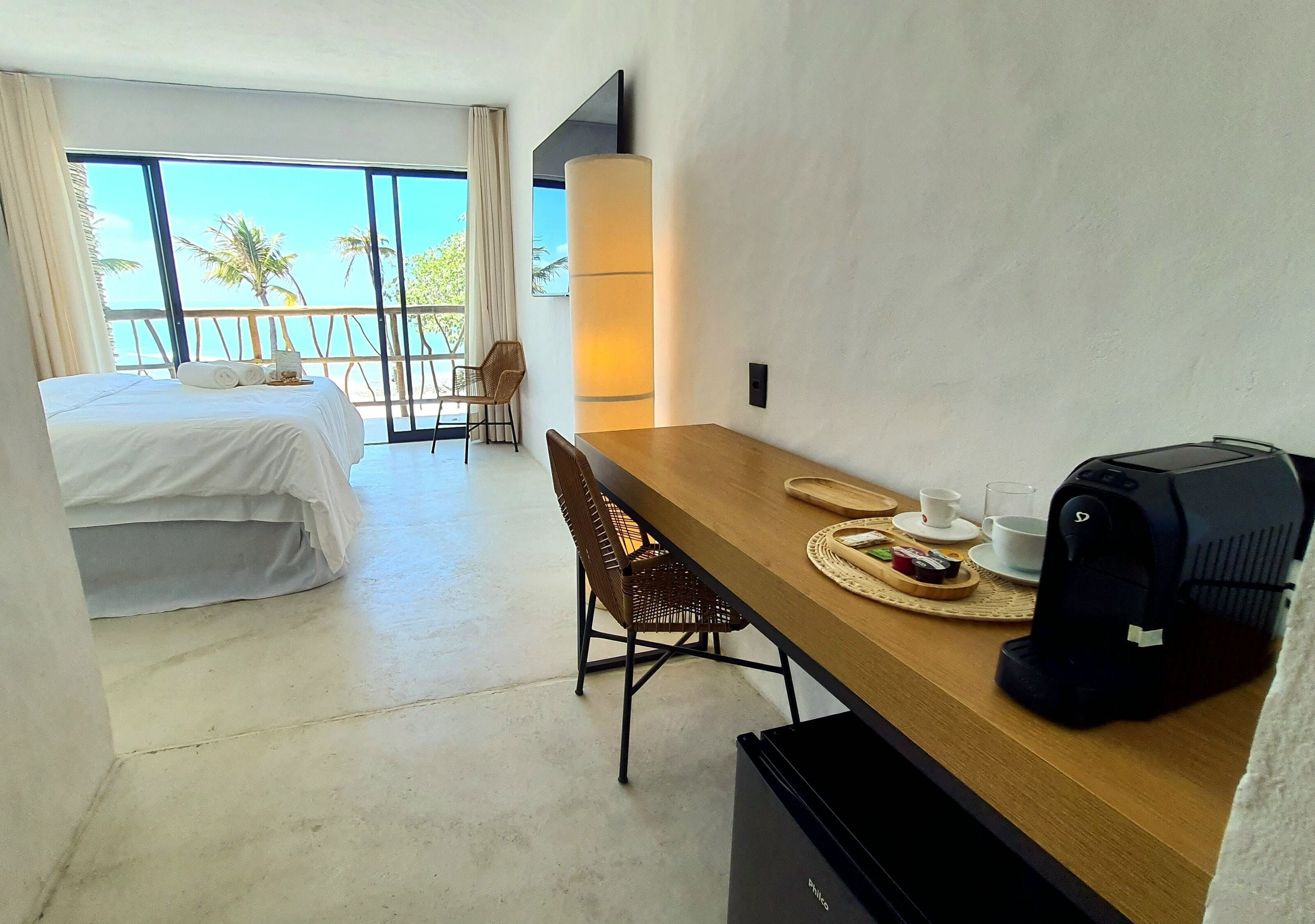 A hotel room with a bed, a dining table with chairs, a coffee machine, and a large window with a view of palm trees.