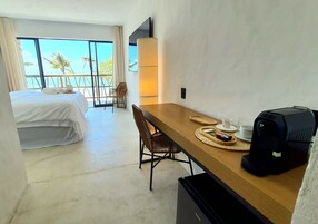Habitación de hotel con cama, una mesa de comedor con sillas, cafetera y un ventanal con vistas a palmeras.