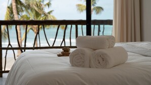Una cama con sábanas blancas y toallas enrolladas, una bandeja de madera con una piedra, y vista a la playa y palmeras a través de la barandilla de un balcón.