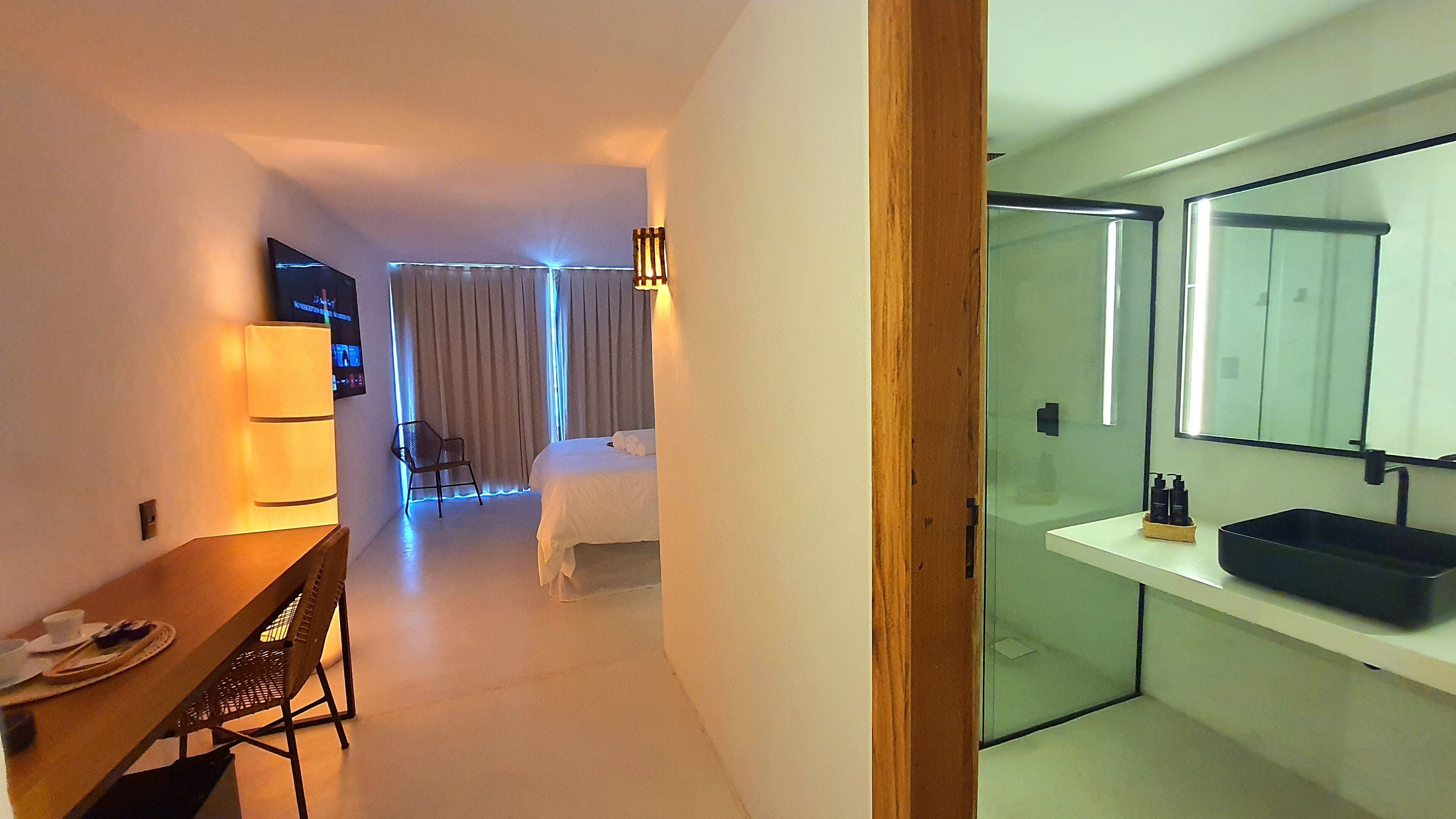 A modern hotel room with a dining area, a bed, and a bathroom with a large mirror and sink.