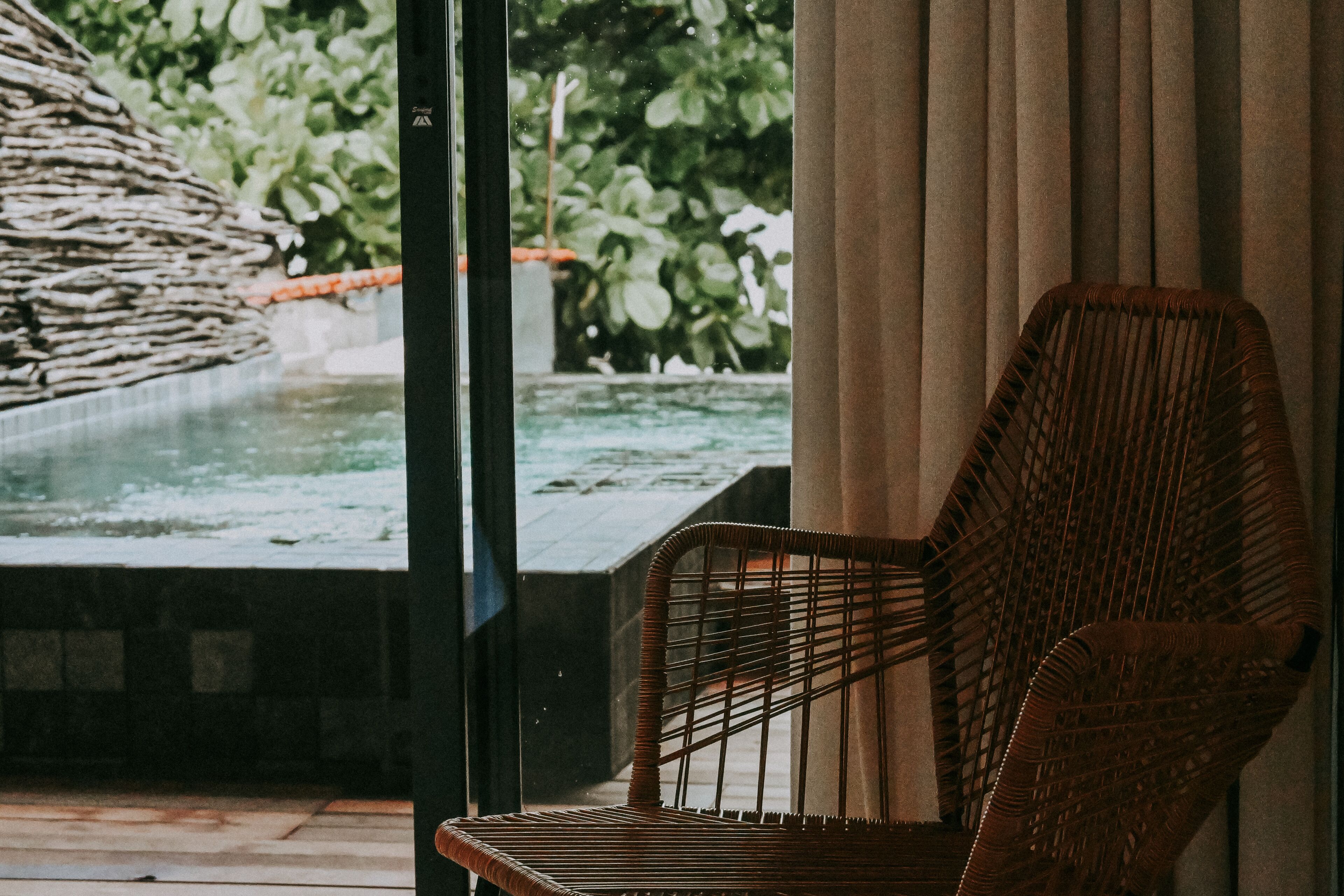 A wicker chair in a room with a view of a water feature and greenery outside.
