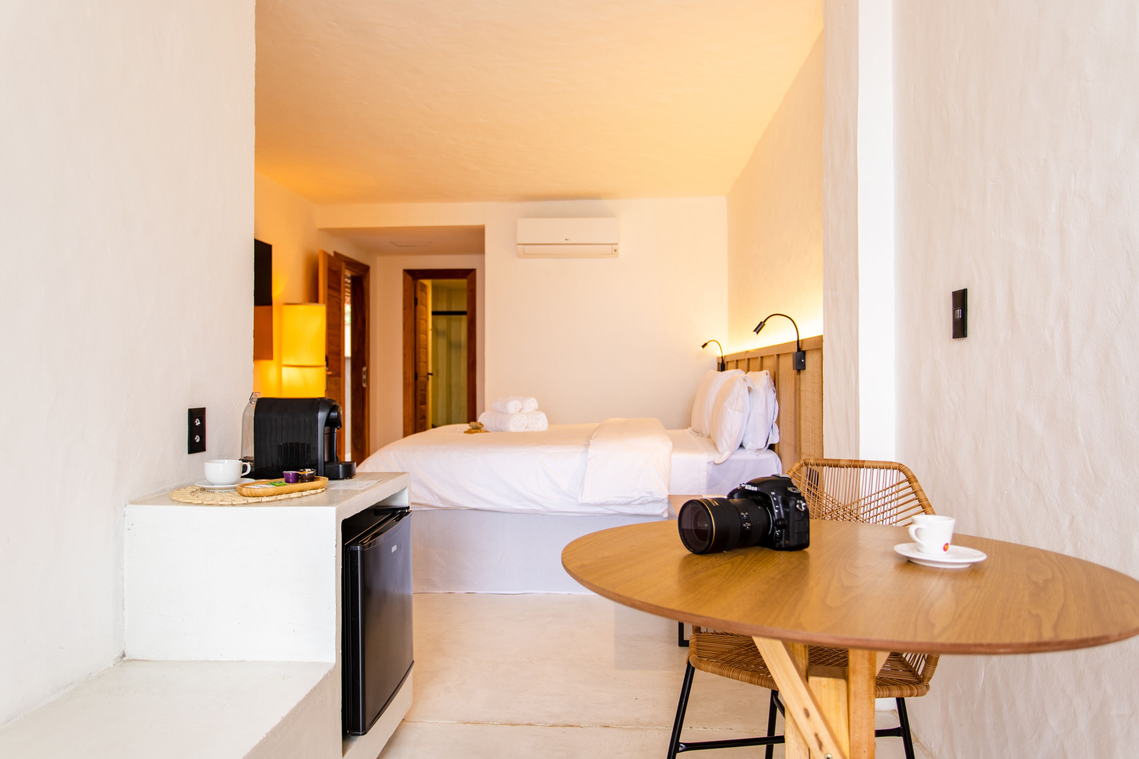 A hotel room with a bed, a dining table with a camera, a coffee maker, and a lamp.