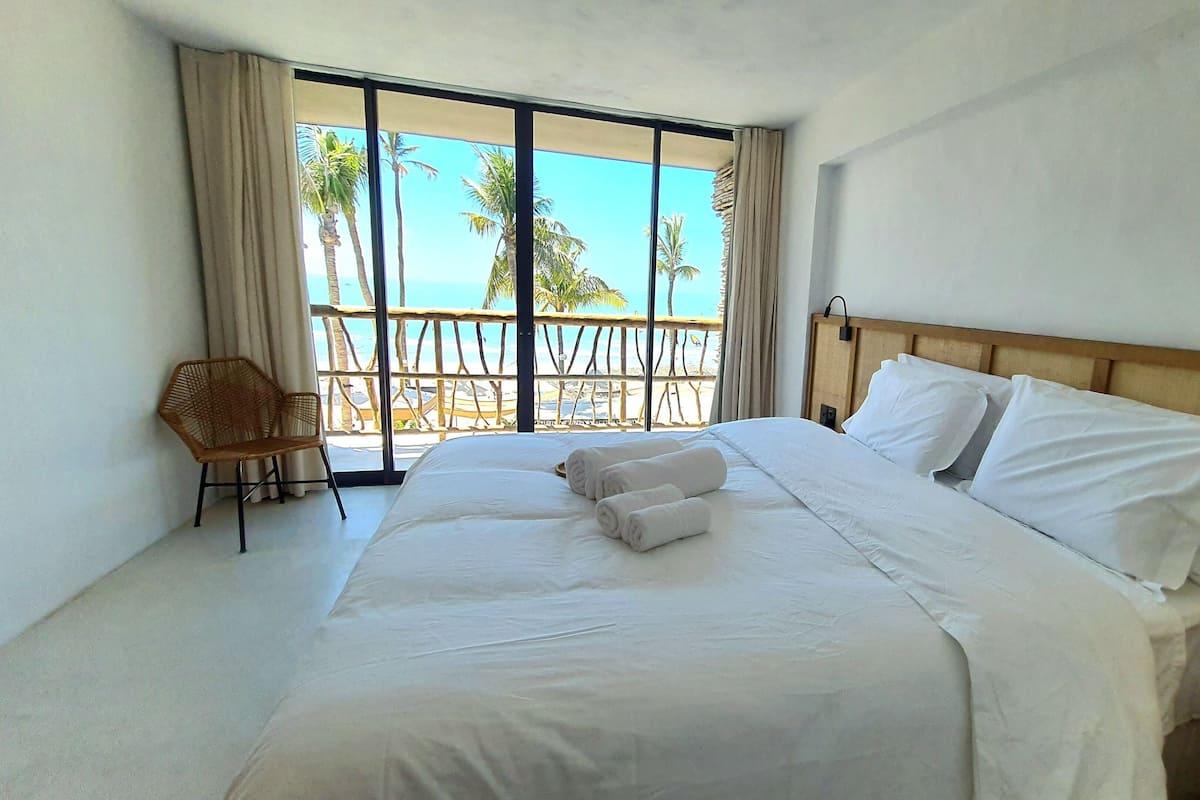 A hotel room with a large bed, a chair, and a view of the beach through a sliding door.