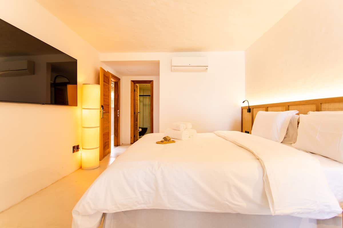 A hotel room with a large bed, white linens, a bedside lamp, and an air conditioning unit on the wall.
