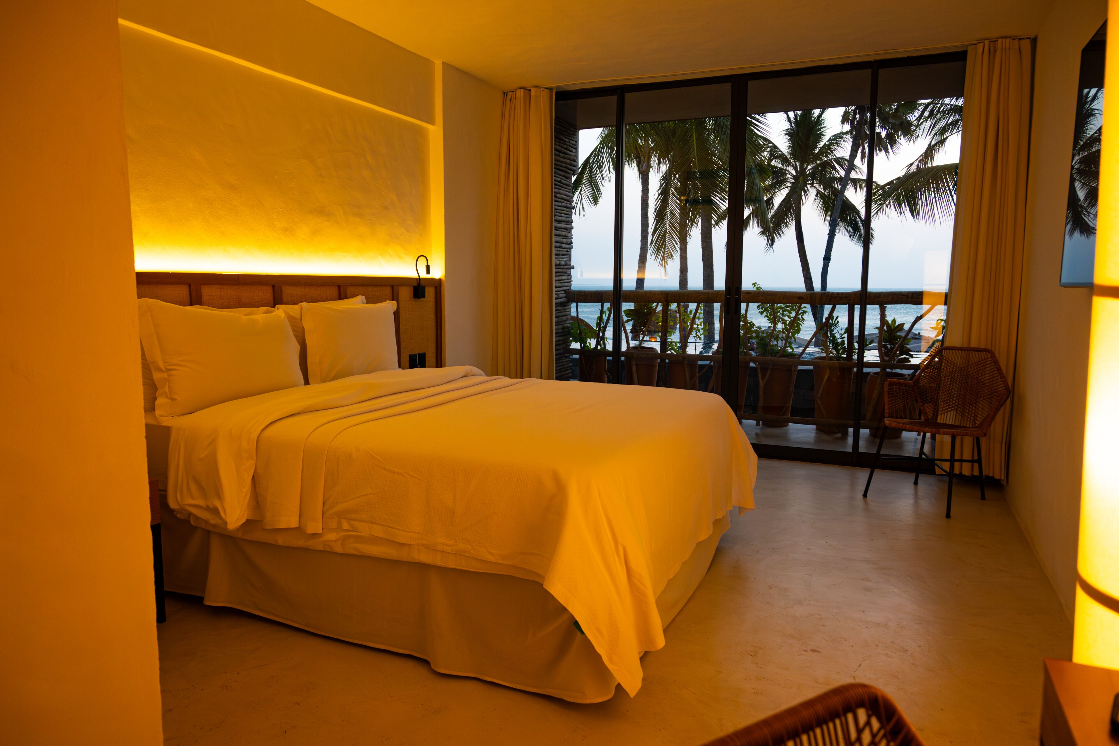 Habitación de hotel con una cama grande, vista a palmeras y al océano desde un balcón, y una silla junto a la ventana.