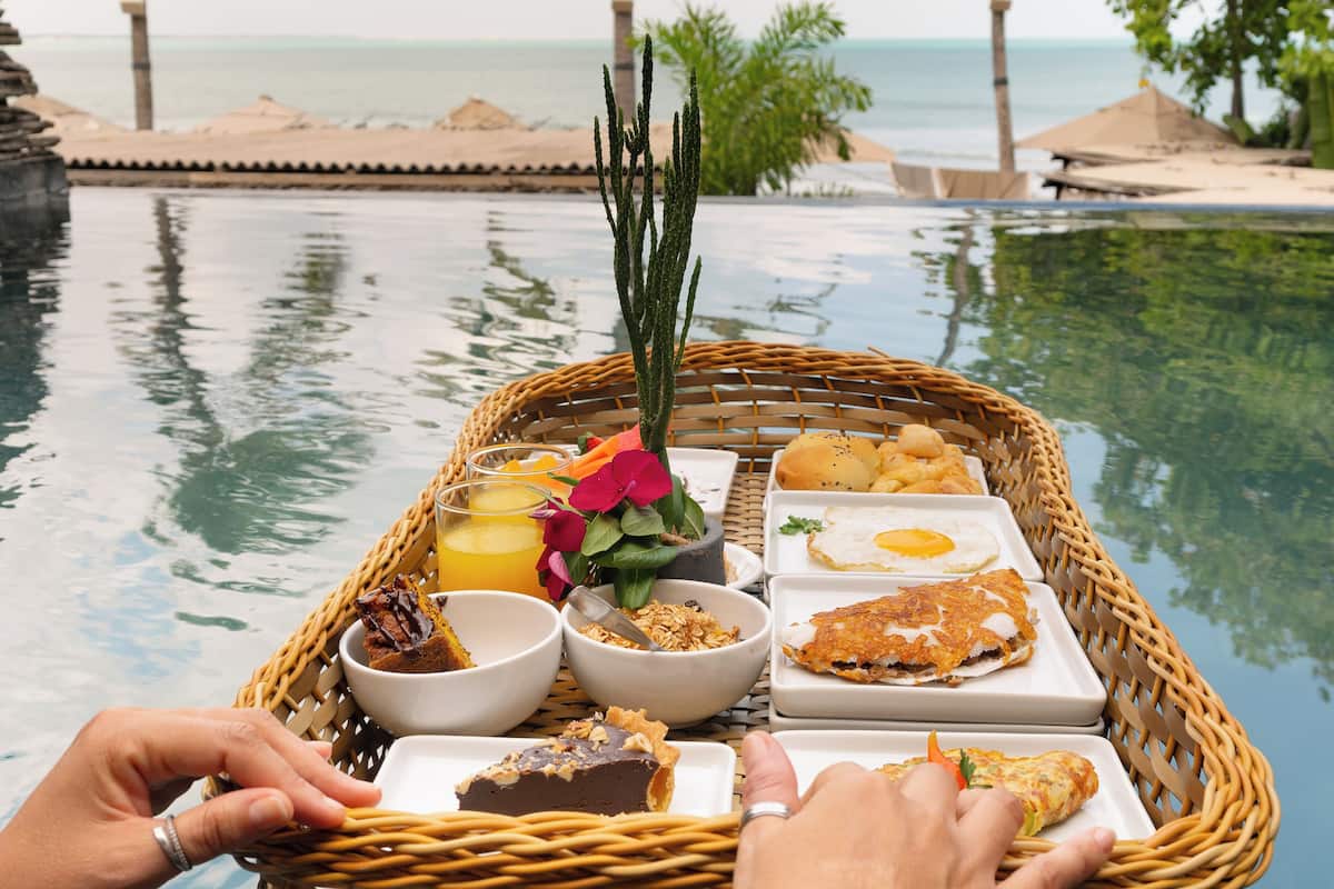 A dining setup by a poolside with a wicker table holding various dishes, including a slice of cake, a bowl of granola, and a plate with a fried egg and sandwich.