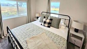 Ett sovrum med en säng, två sängbord med lampor, ett fönster med gardiner och utsikt över husen utanför.