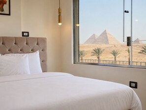 Une chambre d’hôtel avec un lit, une fenêtre donnant sur les pyramides et un luminaire fixé au mur.