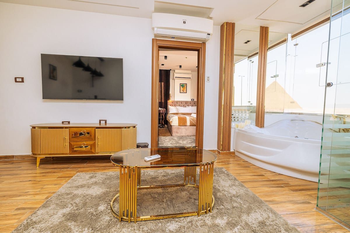 A modern hotel room with a large glass shower enclosure, a round gold-framed table, and a wooden cabinet.