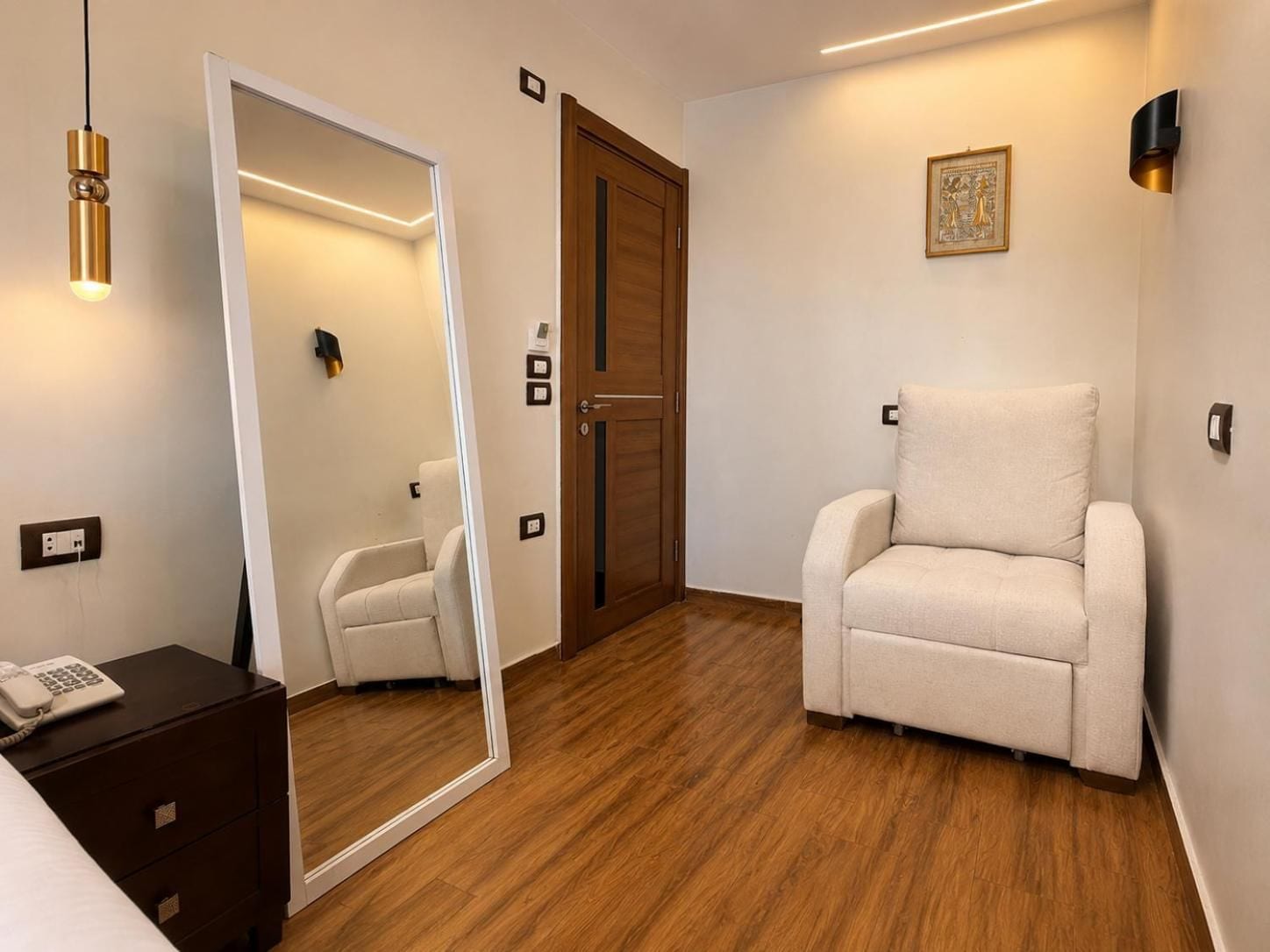 A hotel room with a wooden floor, a white armchair, a bedside table with a phone, a mirror, and a door with a wooden frame.