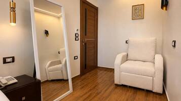 A hotel room with a wooden floor, a white armchair, a bedside table with a phone, a mirror, and a door with a wooden frame.