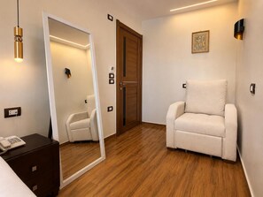 A hotel room with a wooden floor, a white armchair, a bedside table with a phone, a mirror, and a door with a wooden frame. - White Pyramids inn  (Giza)