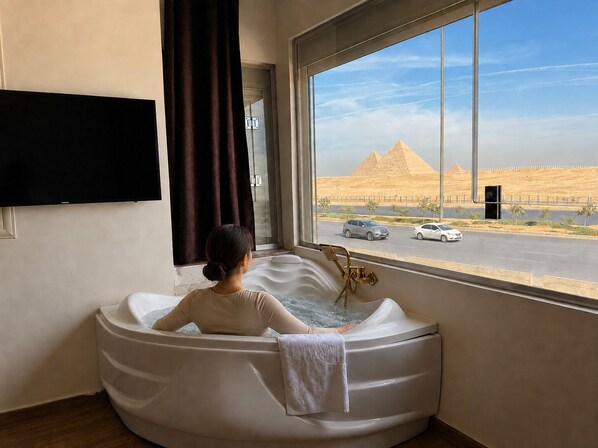 A person relaxing in a bathtub with a view of the pyramids through a large window. - White Pyramids inn  (Giza)