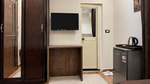 Een hotelkamer met een houten garderobe, een klein houten tafeltje, een aan de muur bevestigde televisie, een koelkast en een deur naar een andere kamer.
