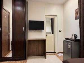 A hotel room with a wooden wardrobe, a small wooden table, a television mounted on the wall, a refrigerator, and a door leading to another room. - White Pyramids inn  (Giza)