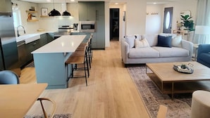 A modern kitchen with a central island, a dining area with a long table, a living room with a sofa and coffee table, and a hallway leading to another room. - Our new lovely home ready for you (Garden City)