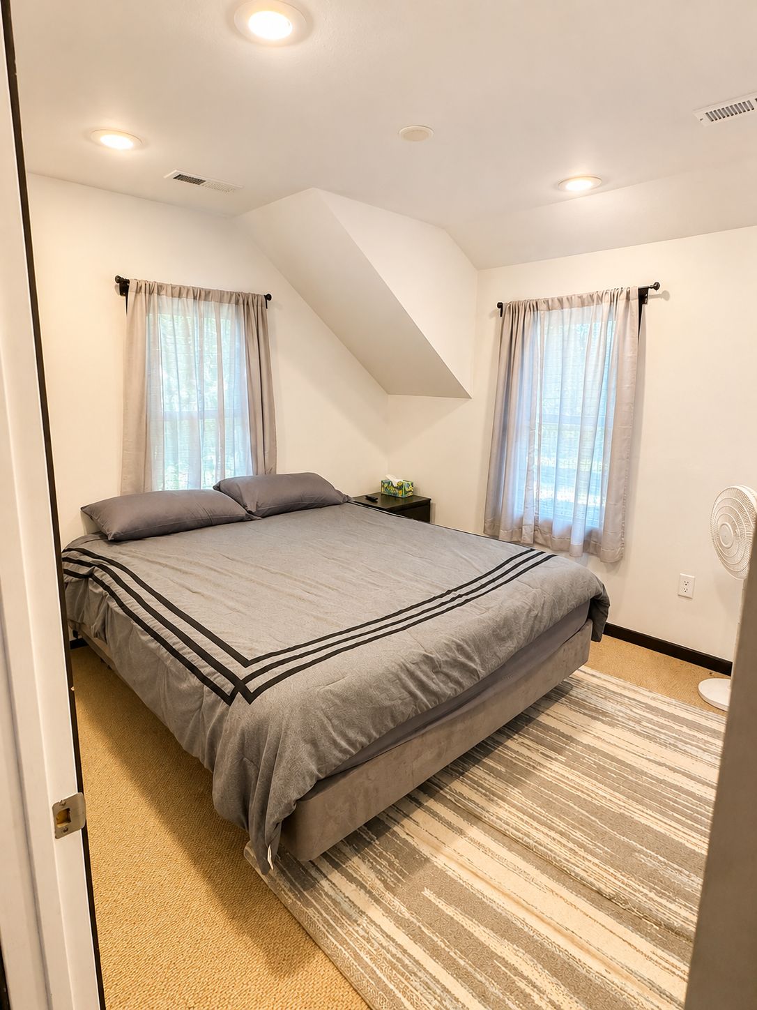 Een slaapkamer met een bed, gordijnen, een nachtkastje, een ventilator en een schuin plafond.