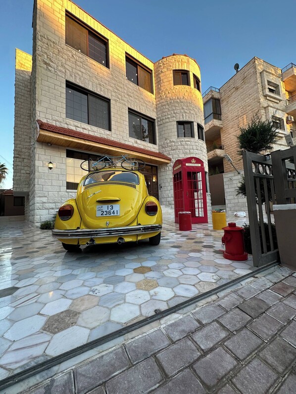 Une voiture jaune, modèle ancien, est garée devant un immeuble à la façade en pierre, à côté d’une cabine téléphonique rouge.