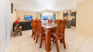 Un comedor con mesa y sillas de madera, un televisor en la pared y una cocina al fondo.