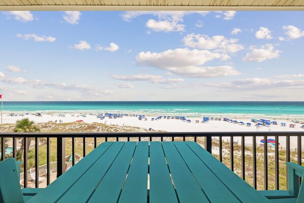 A beach view from a balcony with a table and chairs, a flag, and beach umbrellas. - New Specials - Windancer 404 - Gulf Front, Free Beach Service & Pool (Miramar Beach)