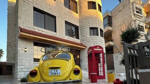 A yellow vintage car parked in front of a building with a stone facade, a red telephone booth, and a gated entrance. - Xx Antica Jabalal-lwebdeh (Amman)