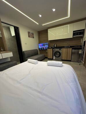 A modern hotel room with a large bed, a flat-screen TV, a washing machine, and a kitchenette.