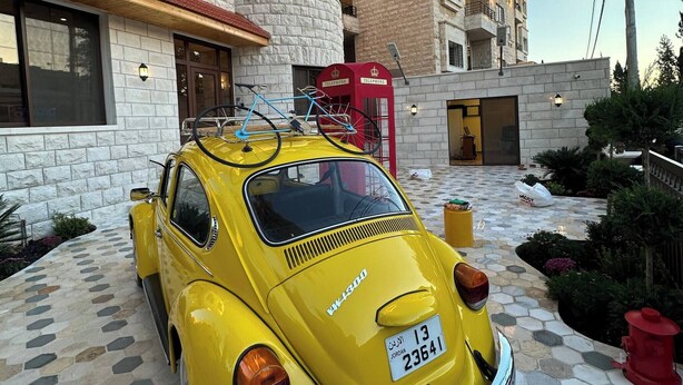 A vintage yellow car parked in front of a building with a stone facade and a red telephone booth. - Xx Antica Jabalal-lwebdeh (Amman)
