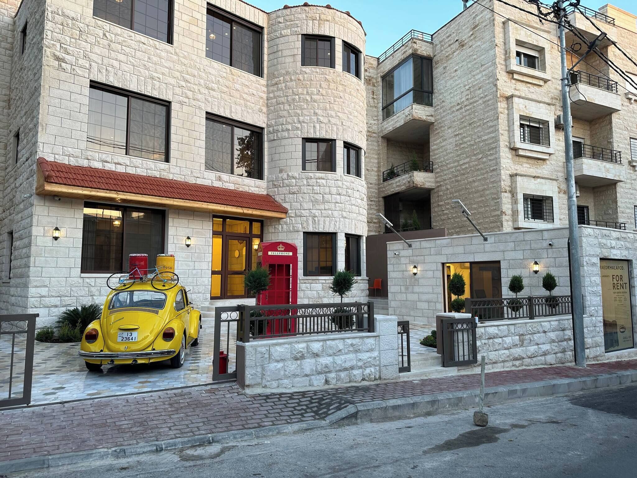 Un immeuble à plusieurs étages avec une façade en pierre, une cabine téléphonique rouge et une voiture jaune garée devant.