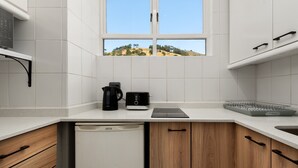 Eine moderne Küche mit weißen Fliesen, einem Geschirrspüler, einer Kaffeemaschine und einem Fenster mit Blick auf Hügel.