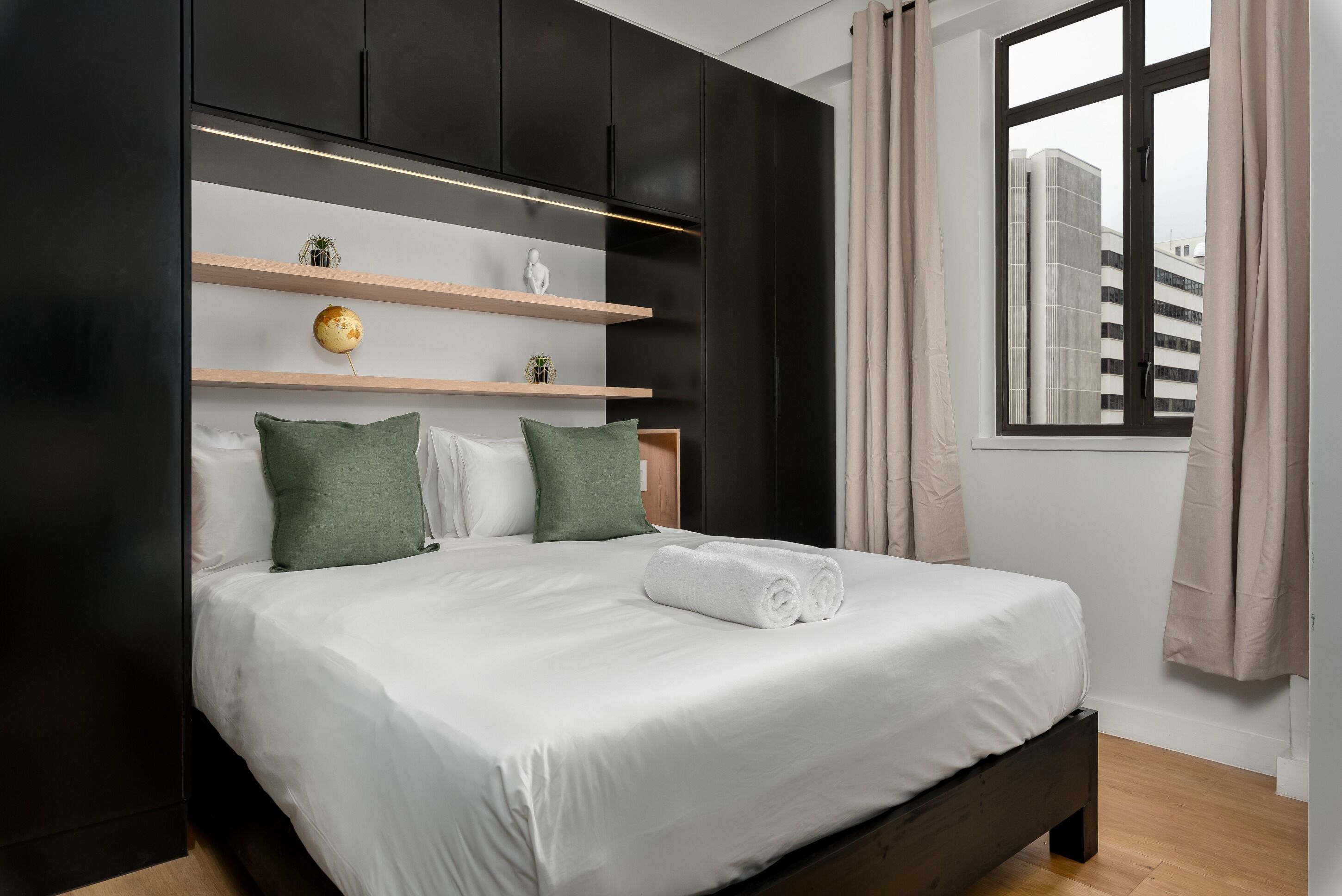 A modern bedroom with a bed, a headboard with shelves, a window with curtains, and a view of buildings outside.
