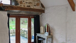 Quarto rústico com parede de pedra, vigas de madeira, uma grande porta de madeira que dá para área externa, um pequeno espelho com toucador e uma cama com manta azul.