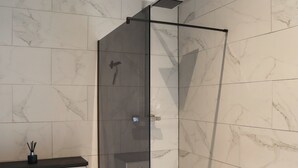 Casa de banho moderna com cabine de duche de vidro, azulejos de mármore e bancada escura.