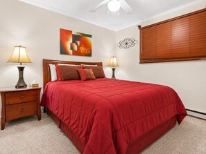Un dormitorio con cama, dos mesitas de noche con lámparas, ventilador de techo, ventana con persianas y una obra de arte en la pared.