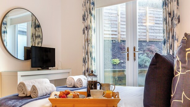 Une chambre d’hôtel avec un lit, un plateau en bois contenant des articles pour le petit-déjeuner, une télévision et un balcon avec des rideaux.