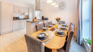 Une cuisine moderne avec des meubles blancs, un îlot central et un coin repas agrémenté d’une longue table pouvant accueillir six personnes.