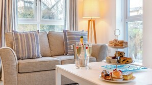 Un salon chaleureux avec un canapé, une table basse sur laquelle sont disposées des pâtisseries, une bouteille de champagne et une lampe.