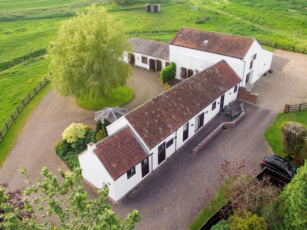 A large white building with a red-tiled roof, surrounded by green fields and a tree.