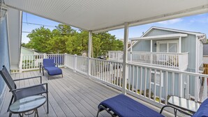 A covered outdoor deck with two lounge chairs and a table, overlooking a residential area with a light blue house and a white fence.